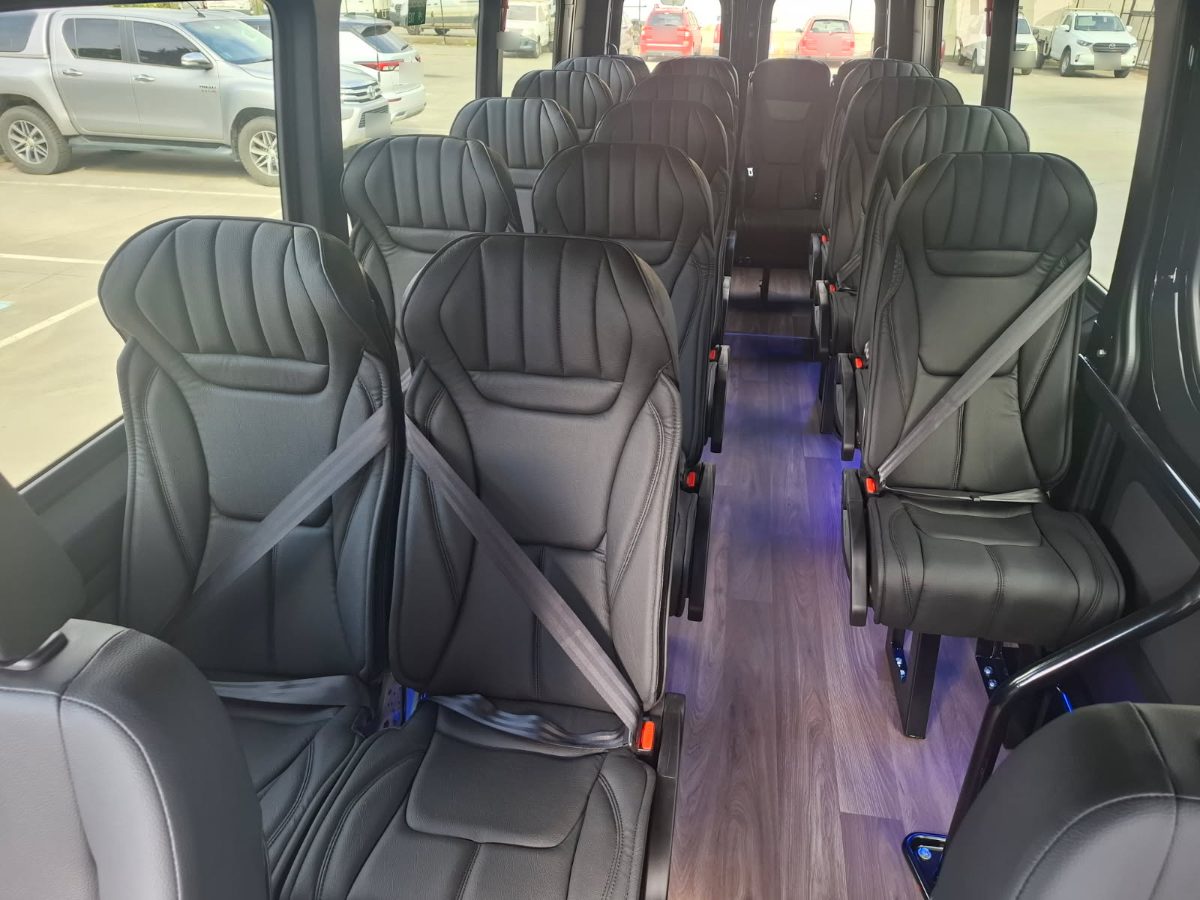 Inside our Mercedes Sprinter 18-Seater: black leather seats with seat belts either side of a lit wood-look aisle