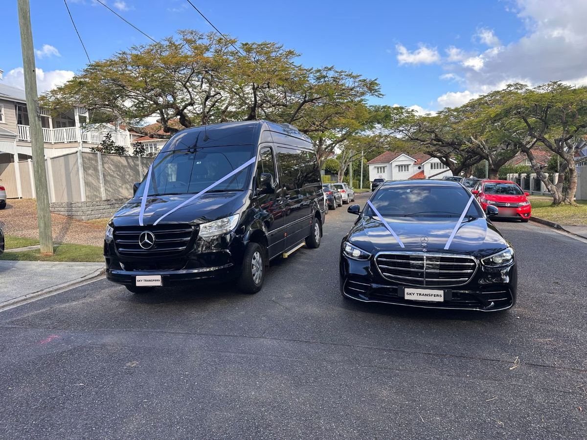 A black Mercedes Sprinter and a black Mercedes S-Class, both dressed with white wedding ribbons, waiting on a tree-lined Brisbane street.